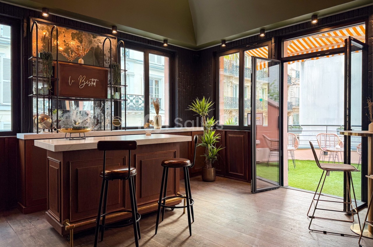 Intérieur d'un bar élégant avec comptoir en bois, tabourets, plantes et terrasse extérieure avec mobilier de jardin.