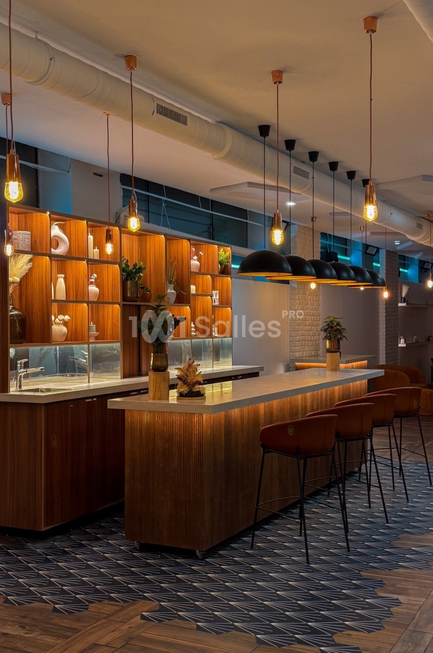 Bar moderne avec comptoir en bois, tabourets, étagères éclairées et suspensions lumineuses dans un espace chaleureux.
