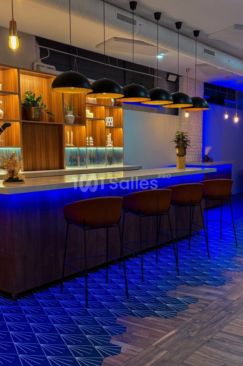 Bar moderne avec comptoir en bois, éclairage suspendu, tabourets en cuir et sol aux motifs géométriques bleus.