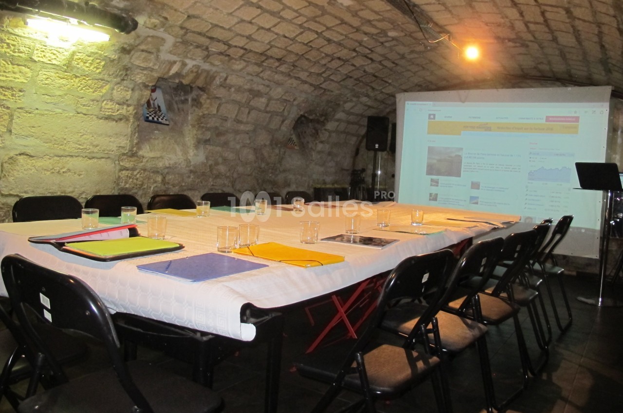 Salle voûtée avec une grande table rectangulaire dressée pour une réunion, équipée d'un projecteur et de chaises noires.