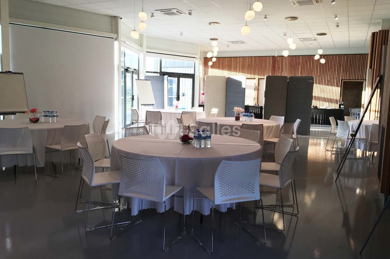 Salle lumineuse aménagée avec des tables rondes, des chaises blanches et des équipements pour une réunion ou un événement.