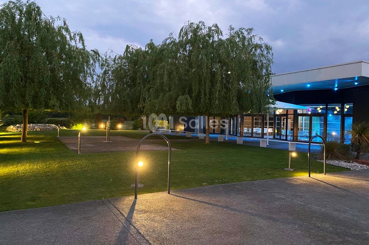 Extérieur d'un bâtiment moderne éclairé, entouré d'arbres et d'une pelouse, avec des lampadaires allumés au crépuscule.