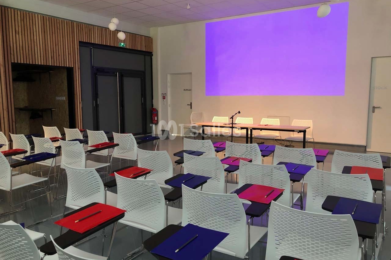 Salle de conférence avec des rangées de chaises blanches équipées de tablettes, face à un écran de projection violet.