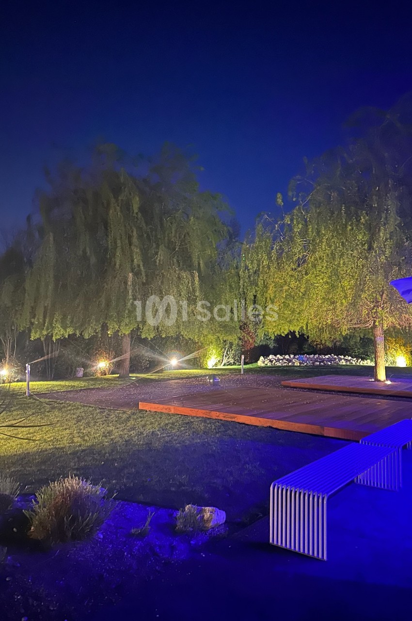 Jardin éclairé de nuit avec arbres, pelouse, terrasse en bois et bancs illuminés par des lumières tamisées.