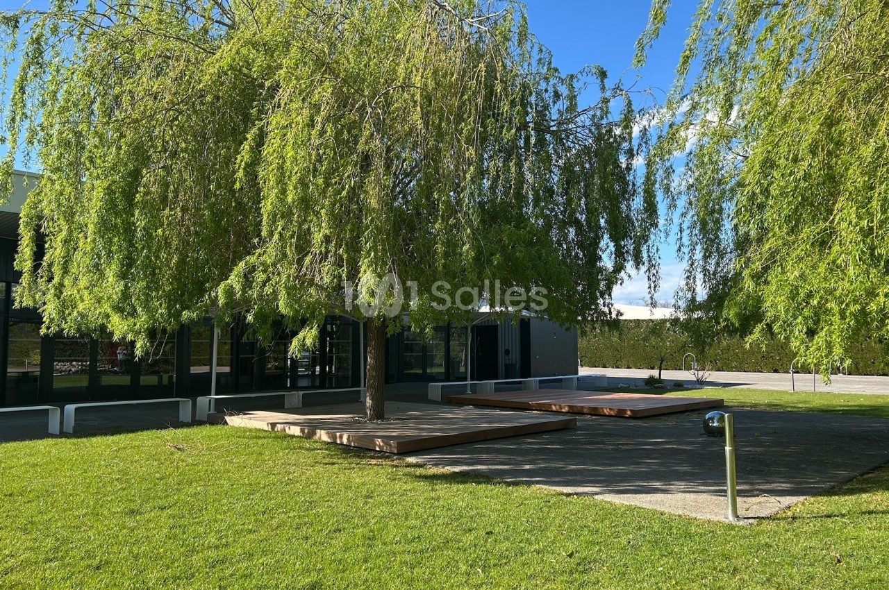 Un espace extérieur avec pelouse, arbres et terrasses en bois, près d'un bâtiment moderne sous un ciel dégagé.