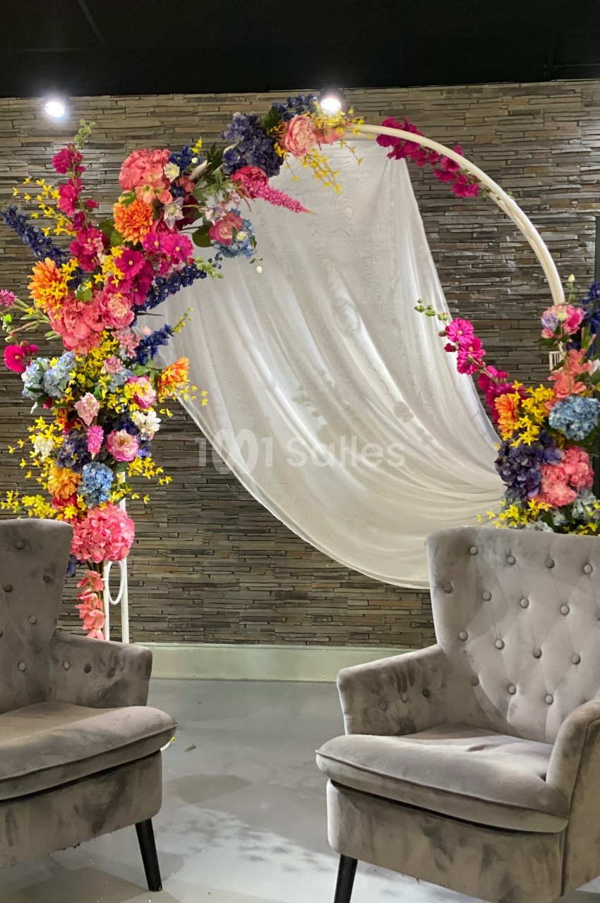 Arche décorative ornée de fleurs colorées et drapée blanche, encadrée par deux fauteuils gris devant un mur texturé.