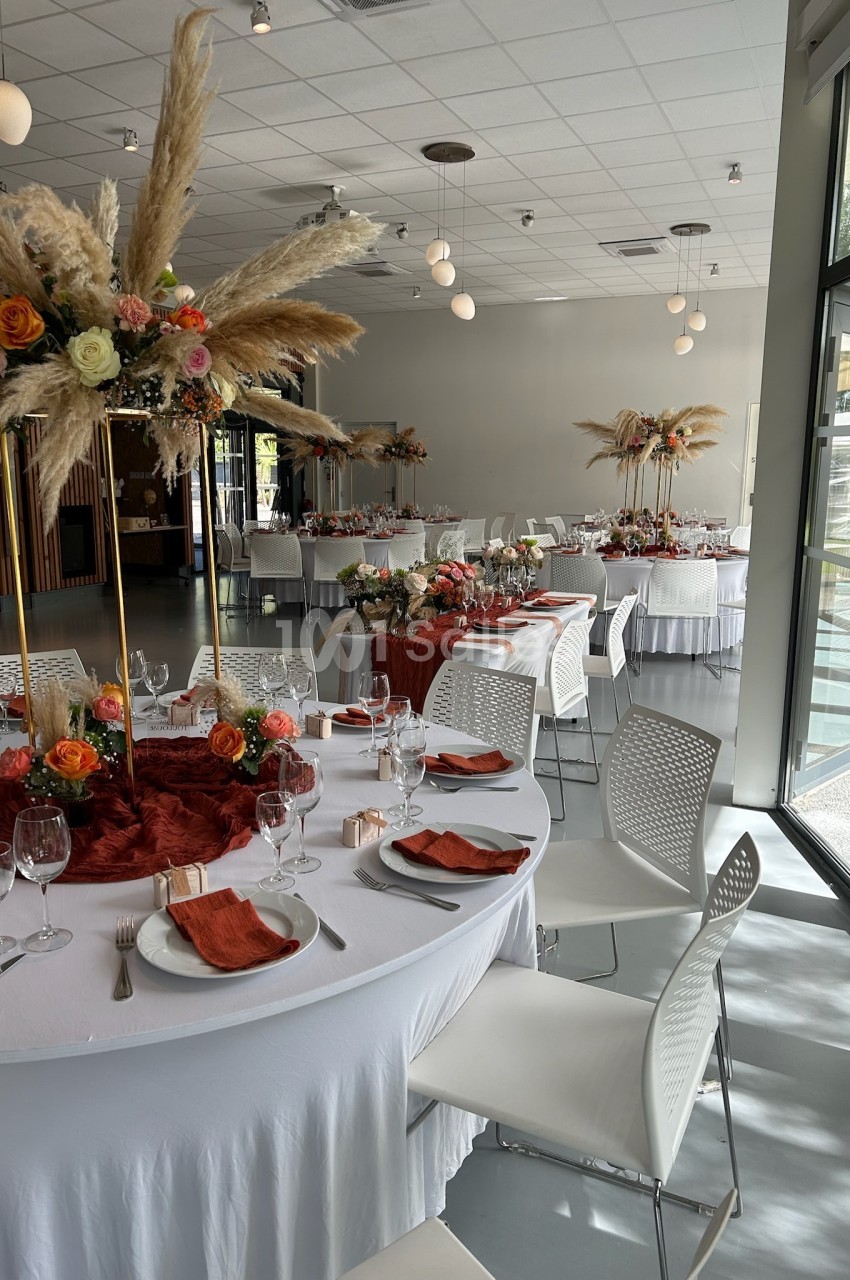 Salle de réception décorée avec des tables rondes, nappes blanches, serviettes orange et centres de table fleuris.