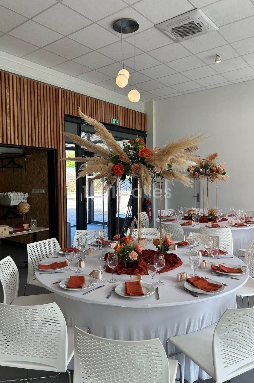 Salle de réception décorée avec des tables rondes, nappes blanches, serviettes orange et centres de table floraux.