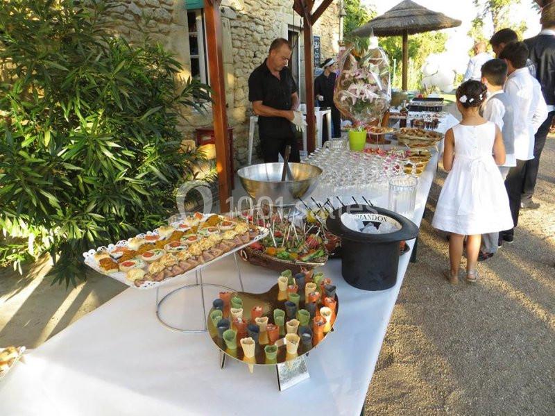 Buffet en extérieur avec verrines, amuse-bouches et boissons, des convives en arrière-plan près d'une table.