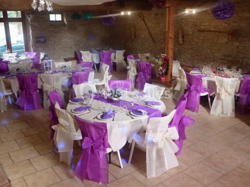 Salle de réception décorée avec des tables rondes, nappes blanches et chaises ornées de nœuds violets et blancs.
