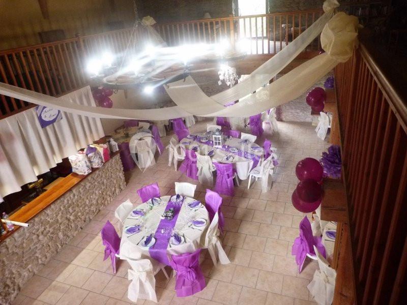 Salle décorée pour un événement, avec tables rondes ornées de nappes violettes et blanches, sous des guirlandes suspendues.