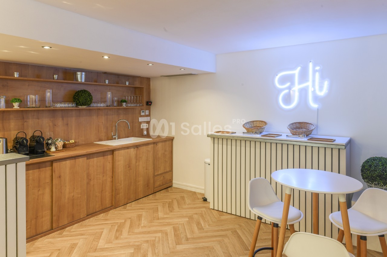 Cuisine moderne avec meubles en bois, sol en parquet, table ronde, et enseigne lumineuse ’Hi’ sur un mur blanc.