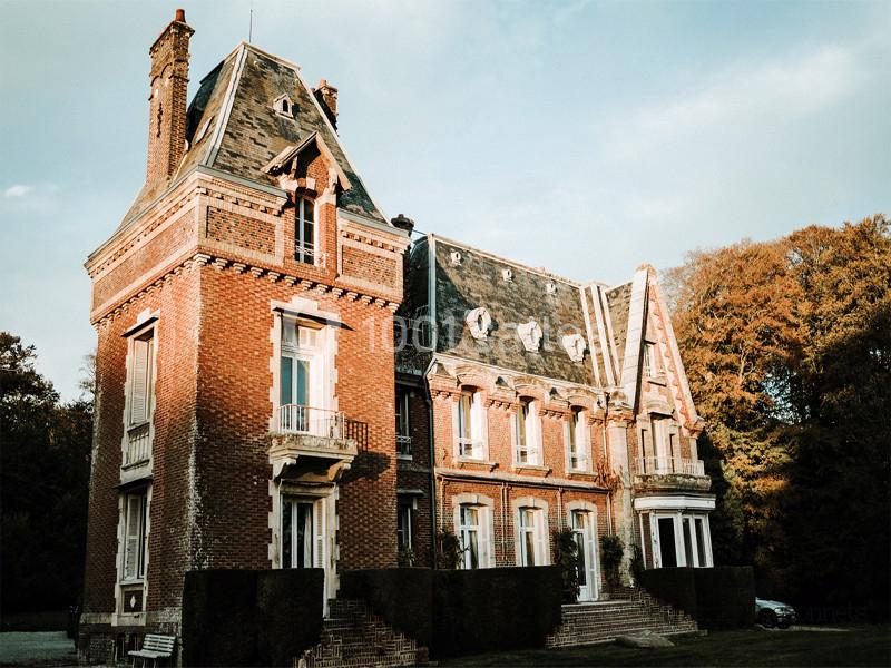 Grande maison en briques rouges avec toit en ardoise, entourée de végétation et baignée par une lumière douce.