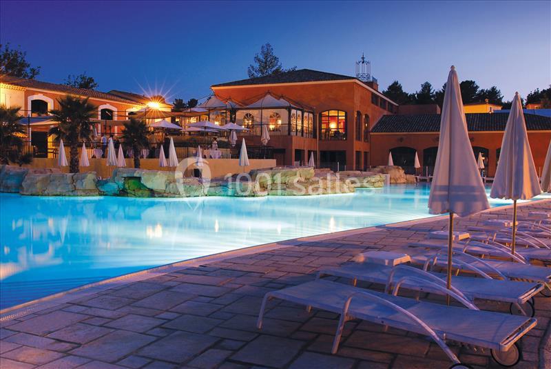 Piscine éclairée en soirée avec transats vides au premier plan et bâtiments en arrière-plan.