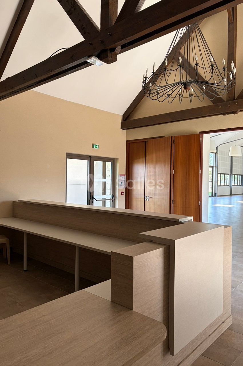 Accueil en bois clair avec comptoirs, situé dans une salle lumineuse aux poutres apparentes et lustre suspendu.