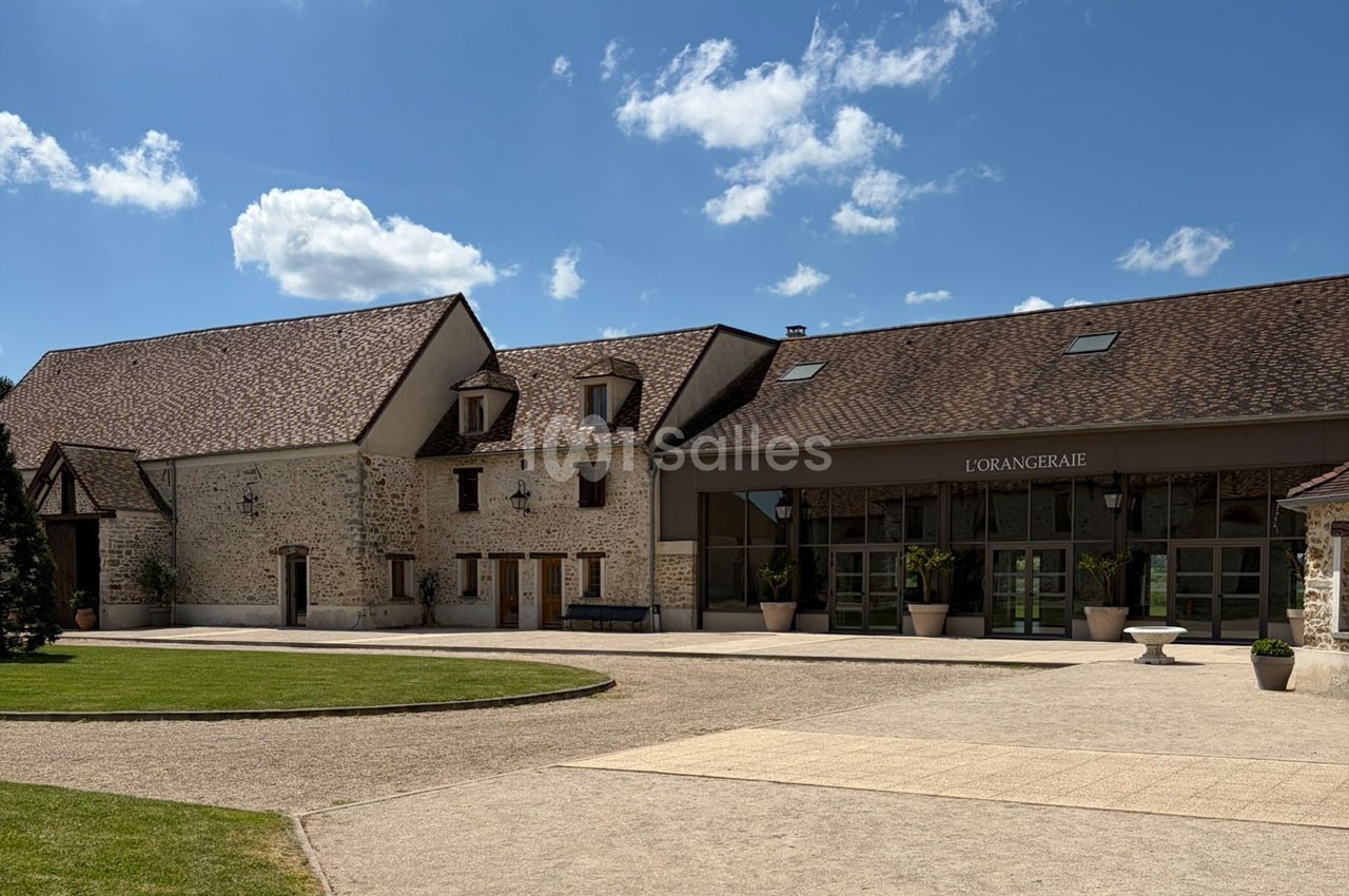 Bâtiment en pierre avec toit en tuiles, grande entrée vitrée et espace extérieur dégagé sous un ciel bleu.