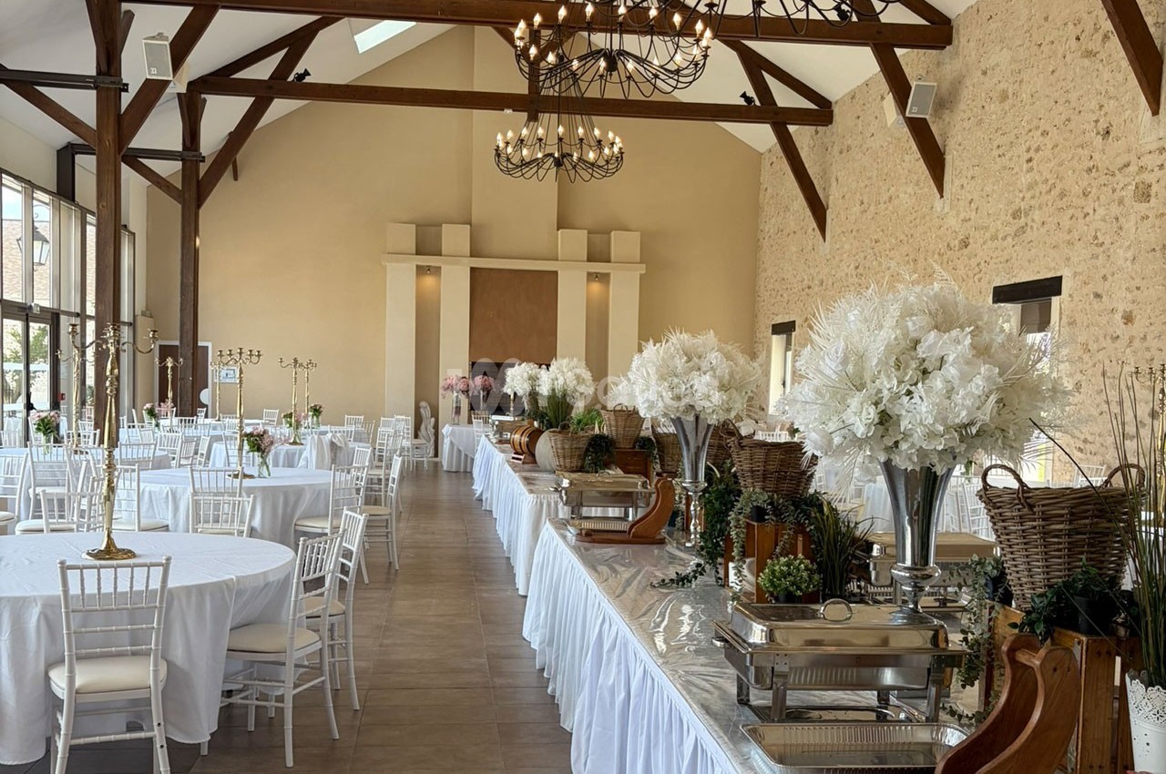 Salle de réception lumineuse avec tables rondes dressées, décorations florales blanches et buffet disposé sur le côté.