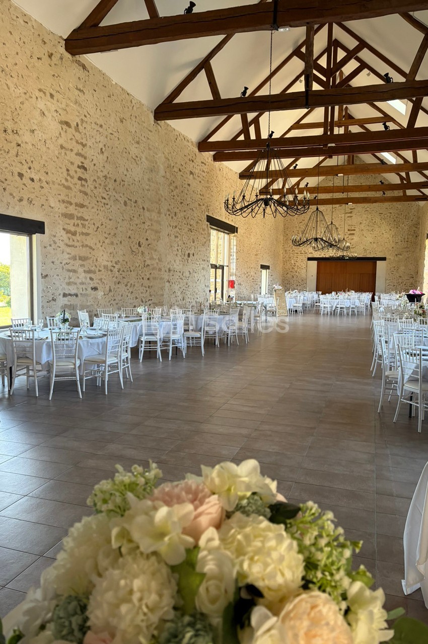 Salle de réception spacieuse avec murs en pierre, poutres apparentes, tables dressées et bouquet de fleurs au premier plan.