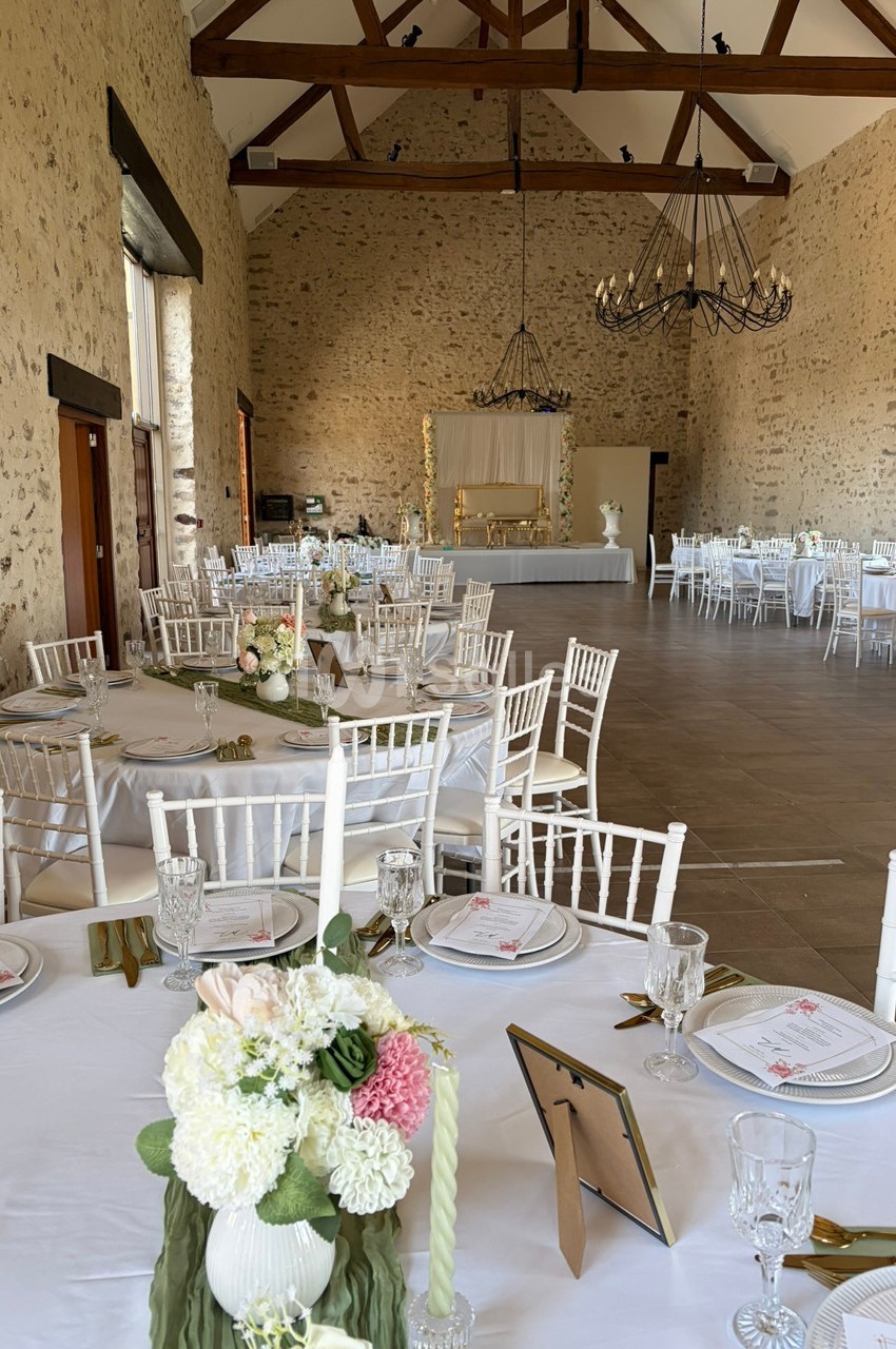 Salle de réception en pierre avec tables dressées, chaises blanches et décorations florales élégantes.