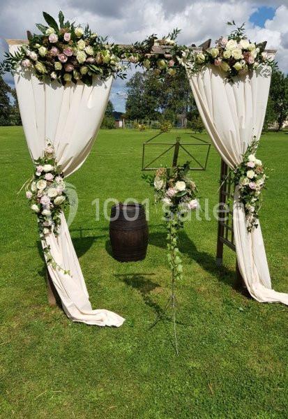 Arche de mariage décorée de fleurs blanches et roses, avec rideaux blancs, sur une pelouse verdoyante.