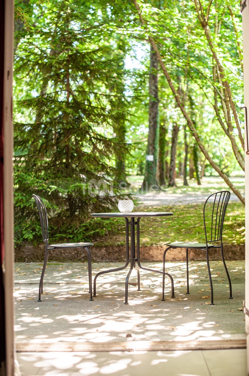 Table et deux chaises en métal noir sur une terrasse ombragée, entourées d'arbres dans un cadre naturel verdoyant.