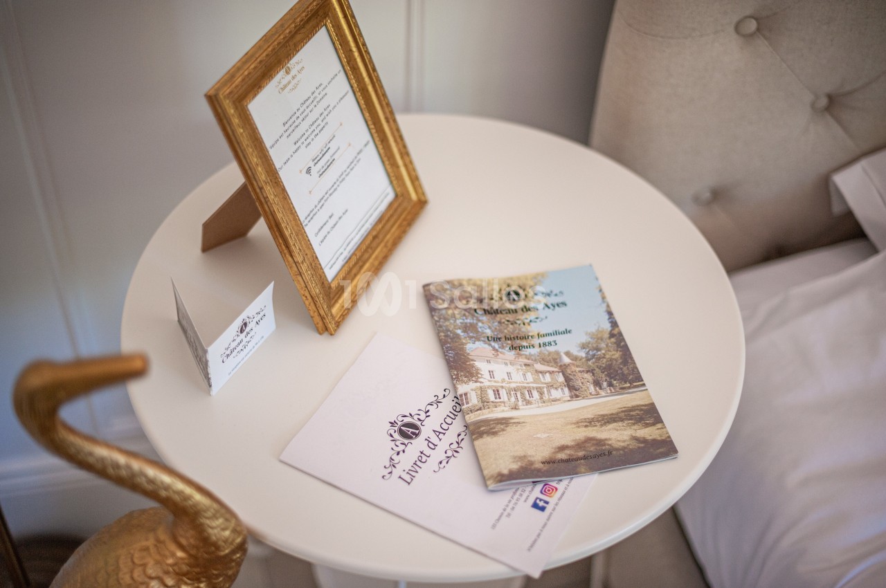 Documents et cadre doré posés sur une table ronde blanche près d'un lit, dans un décor élégant et lumineux.