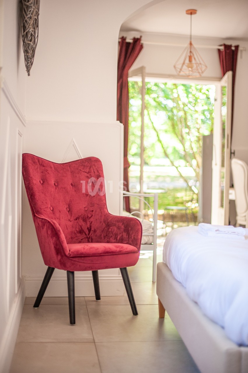 Fauteuil rouge en velours près d'un lit dans une chambre lumineuse avec vue sur un jardin à travers une porte ouverte.