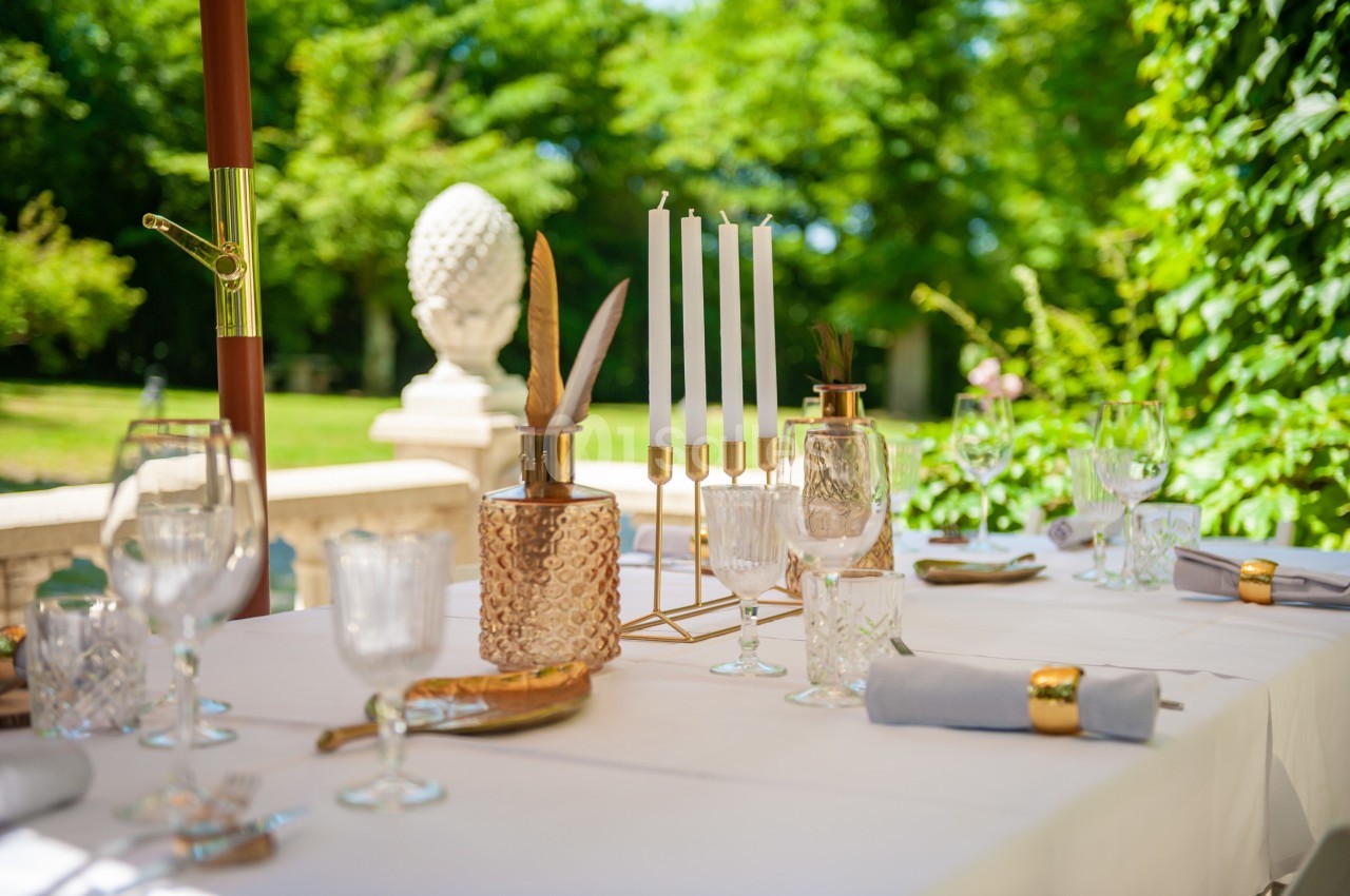 Table élégamment dressée en extérieur avec bougeoirs, verres en cristal et vue sur un jardin verdoyant.