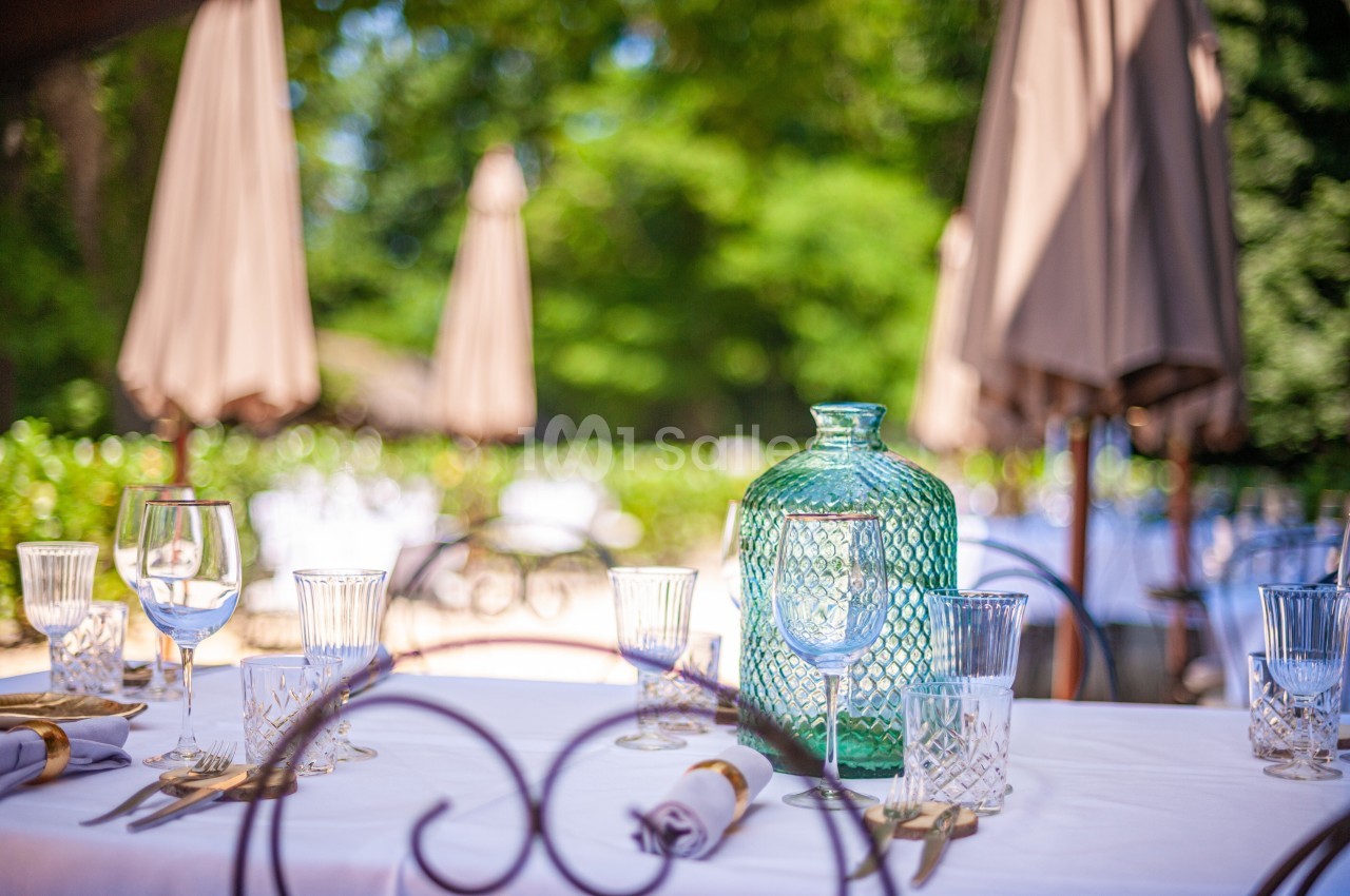 Table dressée en extérieur avec une carafe en verre vert, des verres et des serviettes, sous des parasols beige.