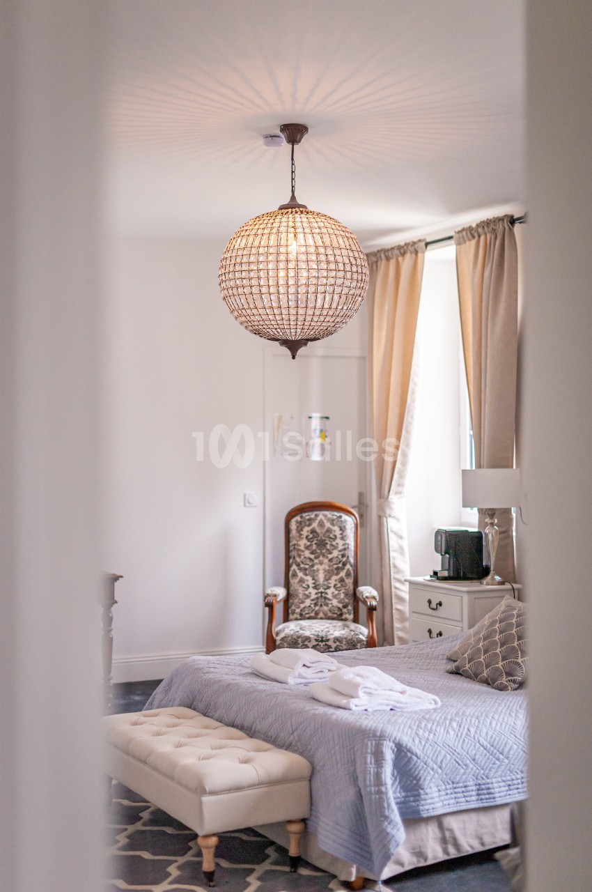 Chambre lumineuse avec un lit, un fauteuil, une commode et un lustre en perles, décorée dans des tons clairs et neutres.