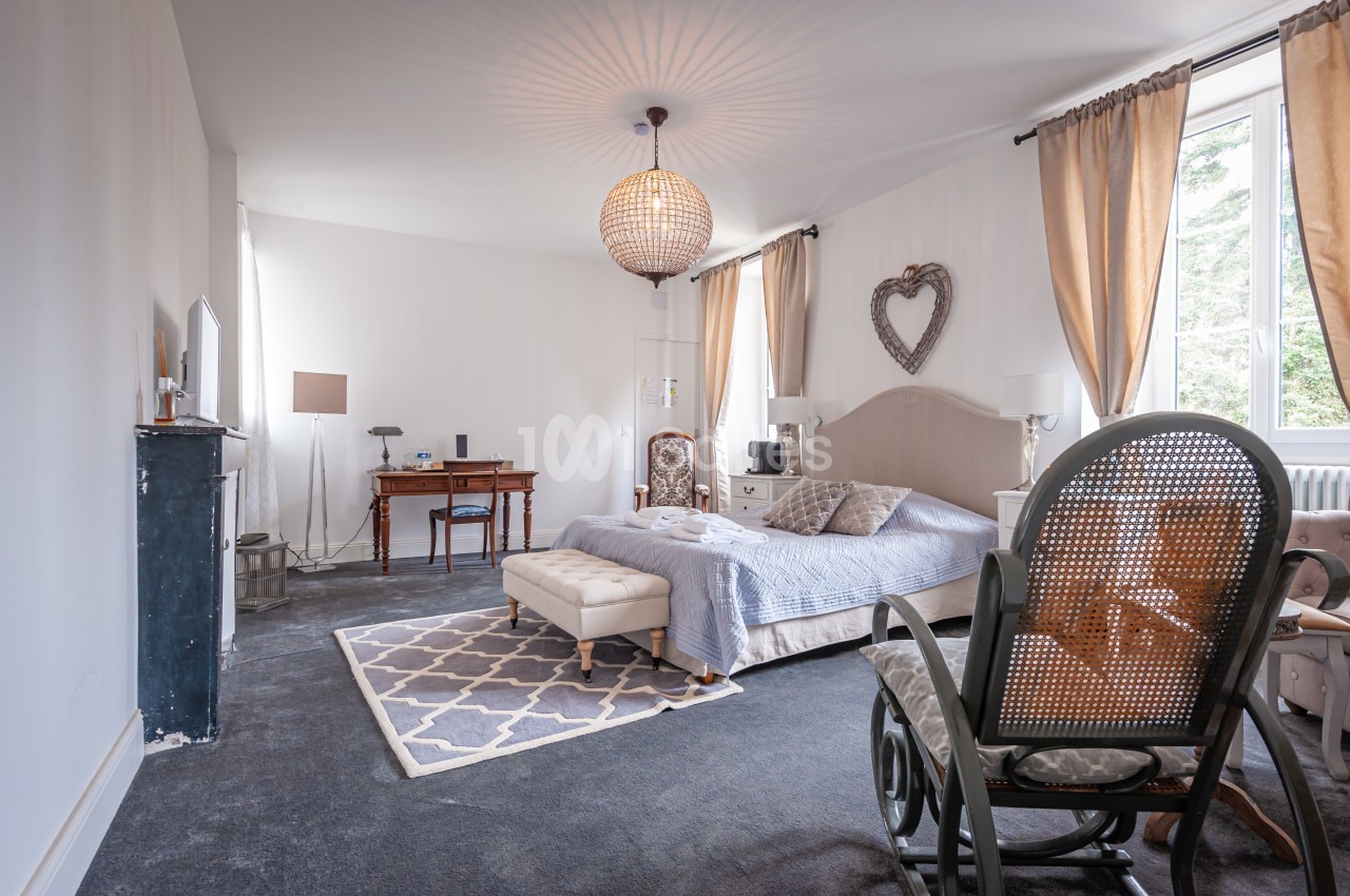 Chambre lumineuse avec lit double, bureau en bois, fauteuil à bascule et grandes fenêtres avec rideaux beiges.