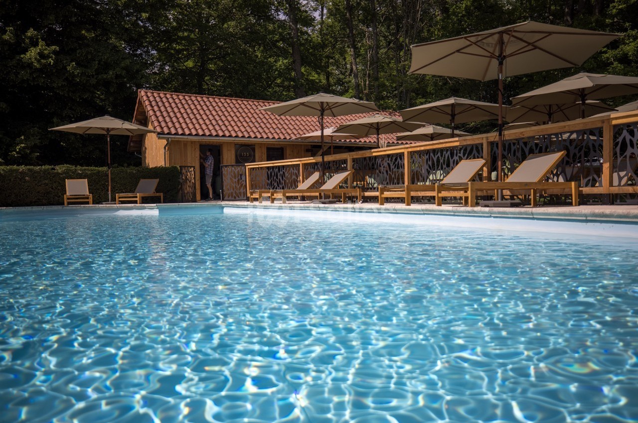 Piscine extérieure entourée de chaises longues, parasols et cabanon en bois, bordée par des arbres.
