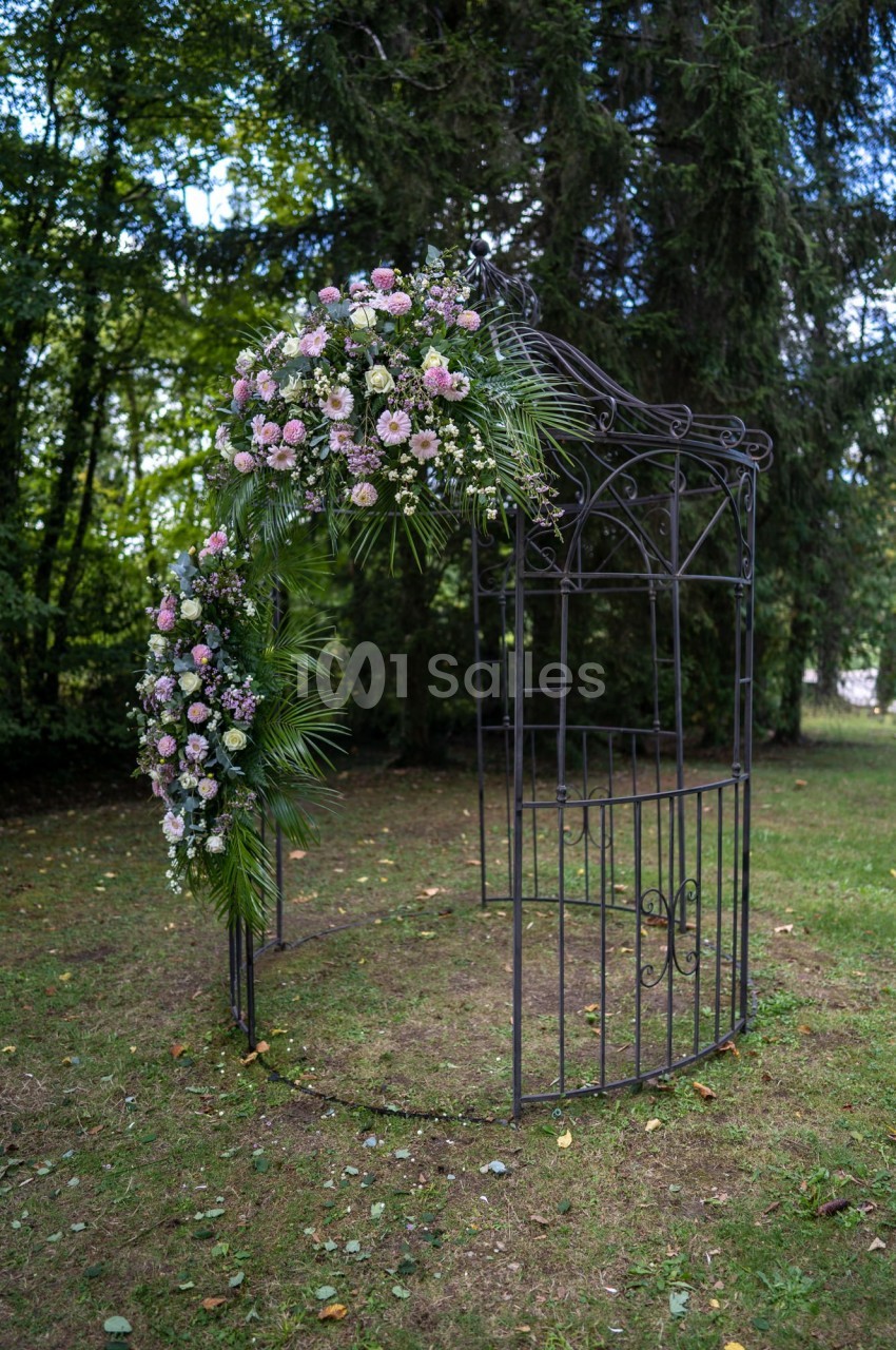Structure métallique décorée de fleurs roses et blanches dans un jardin entouré d'arbres.