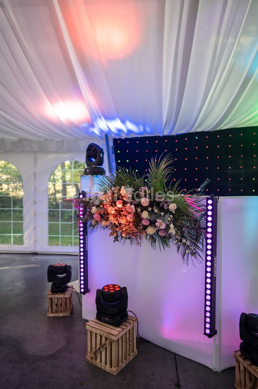 Table de DJ décorée de fleurs et éclairée par des lumières colorées sous une tente blanche.
