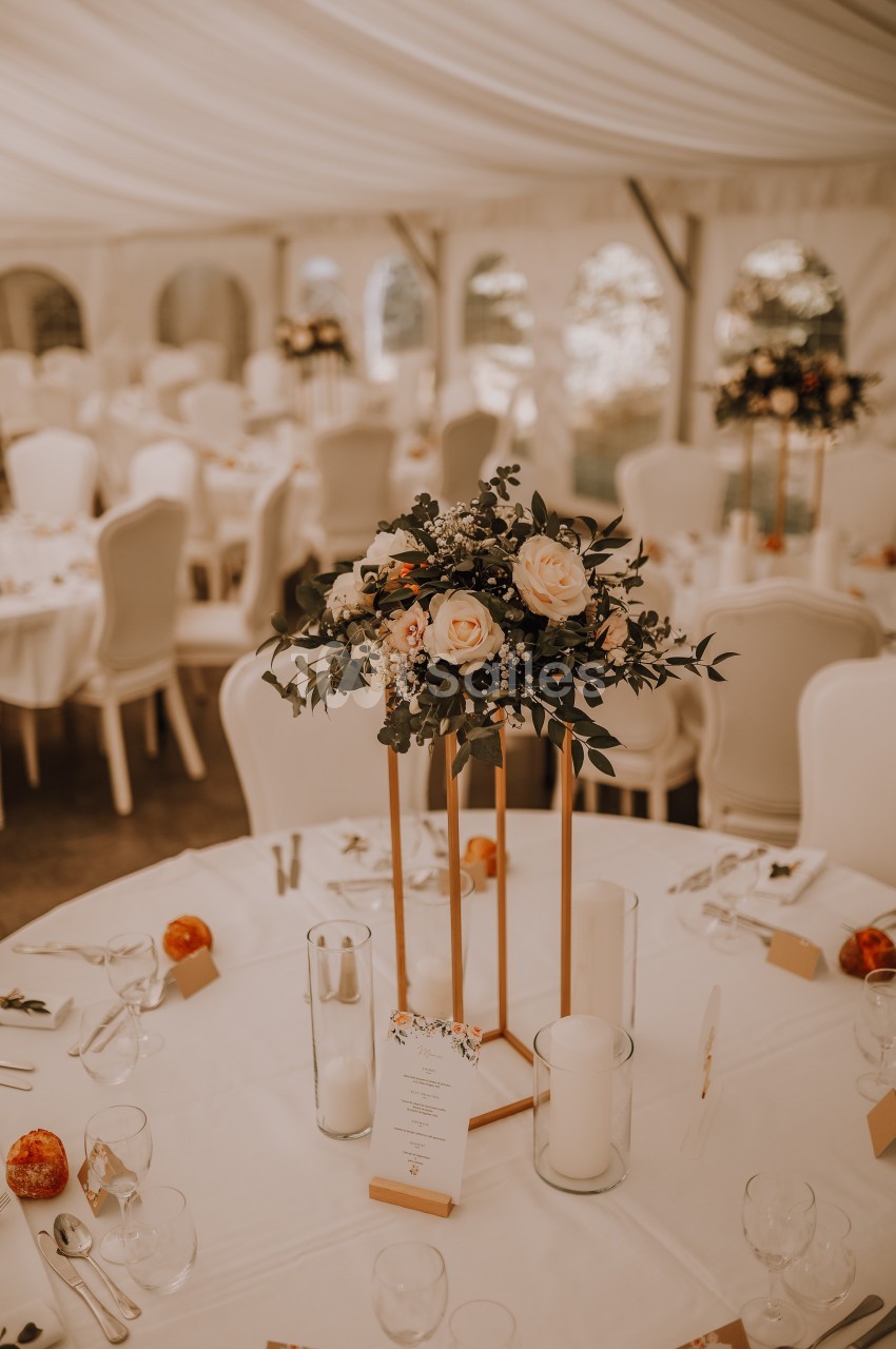 Salle de réception décorée avec des tables rondes, centres de table floraux et chaises blanches sous une tente.