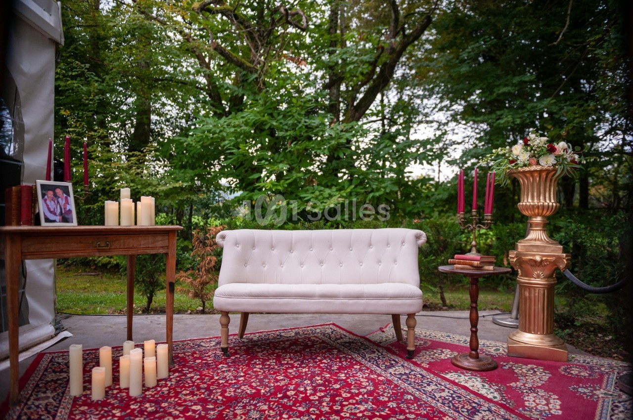 Canapé blanc capitonné sur un tapis rouge, entouré de bougies, d'une table en bois et d'un vase doré dans un jardin.