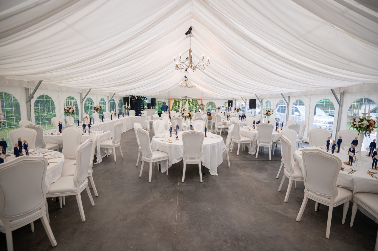 Salle de réception sous une tente blanche avec tables rondes dressées, chaises élégantes et décoration florale.