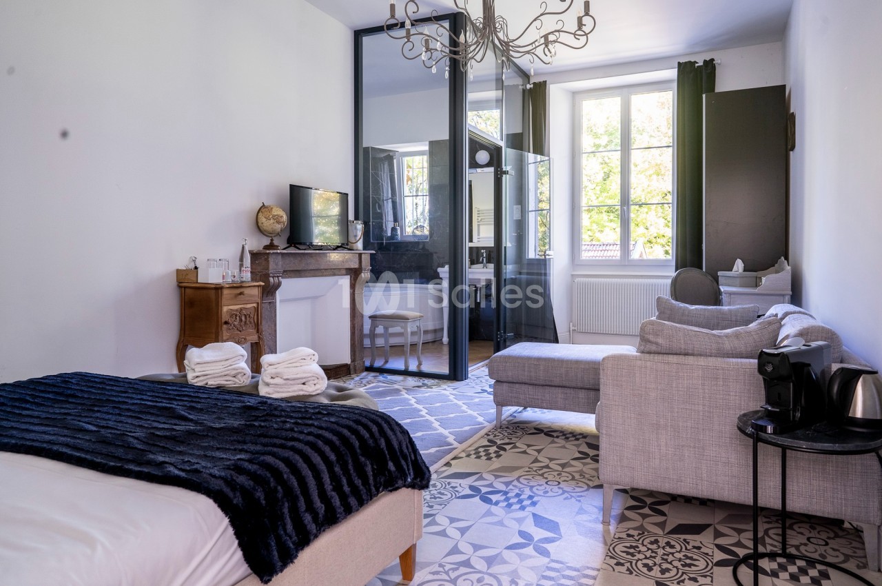 Chambre lumineuse avec lit, canapé, cheminée, bureau et grande fenêtre donnant sur un extérieur verdoyant.