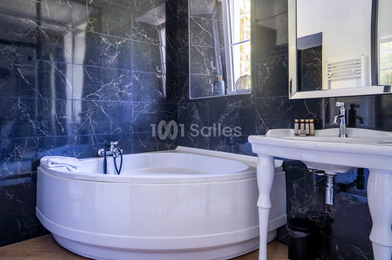 Salle de bain avec baignoire d'angle blanche, murs en marbre noir, lavabo sur pied et fenêtre laissant entrer la lumière.