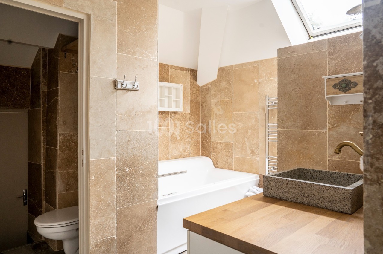 Salle de bain avec baignoire blanche, lavabo en pierre, murs en carrelage beige et lumière naturelle d'une fenêtre.