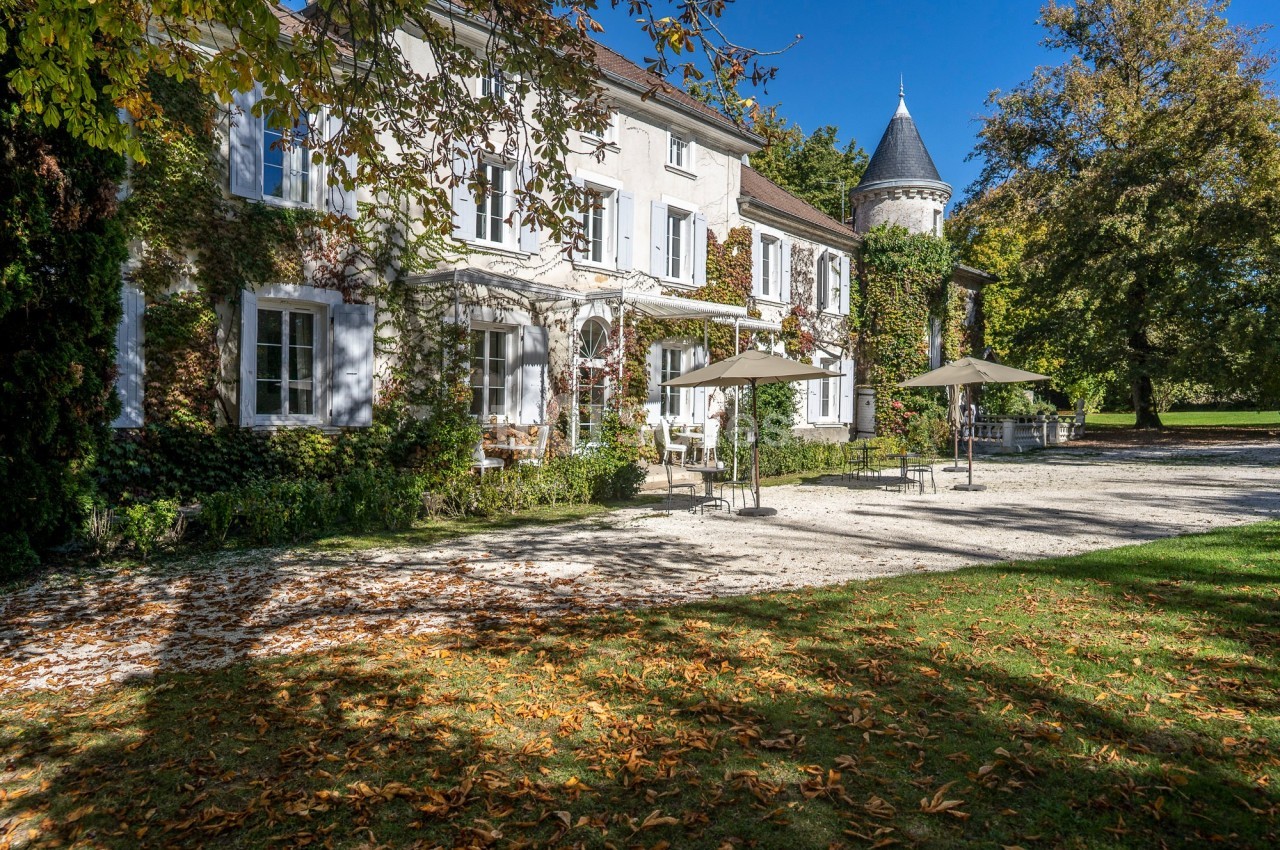 Façade d'une maison de campagne entourée de végétation, avec terrasse ombragée et tour en arrière-plan.