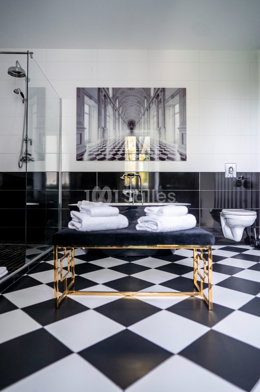 Salle de bain moderne avec carrelage noir et blanc, douche vitrée, banc doré et serviettes pliées.