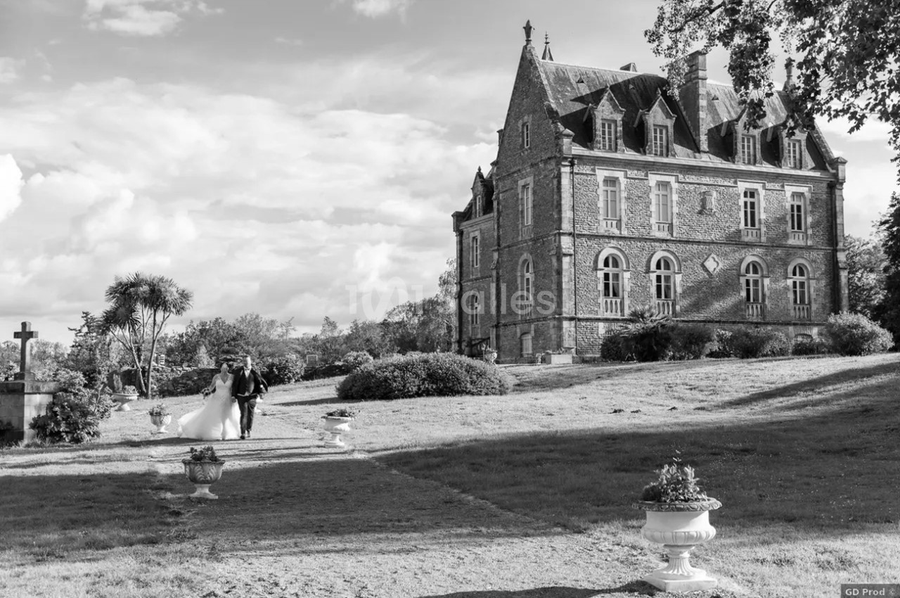 Un couple marche dans un jardin devant un château en pierre sous un ciel partiellement nuageux.