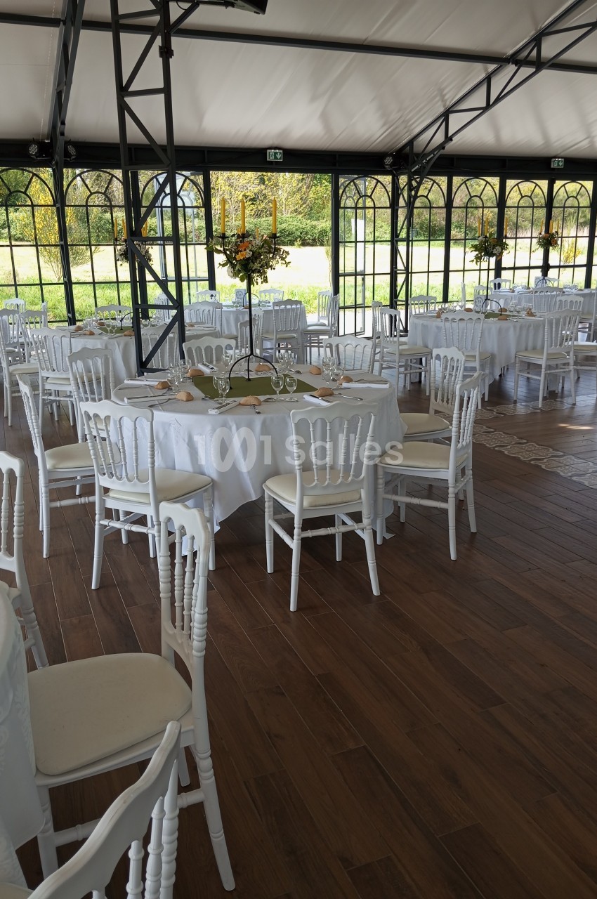 Salle de réception lumineuse avec tables rondes dressées, nappes blanches et chaises blanches, vue sur un jardin.