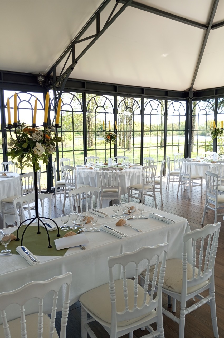 Salle lumineuse avec tables dressées, nappes blanches, chaises en bois, chandeliers et vue sur un jardin verdoyant.