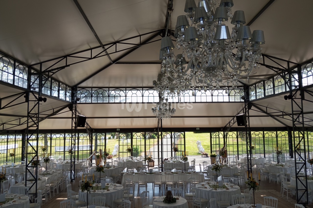 Salle de réception lumineuse avec tables rondes dressées, chaises blanches et lustres suspendus sous une verrière.