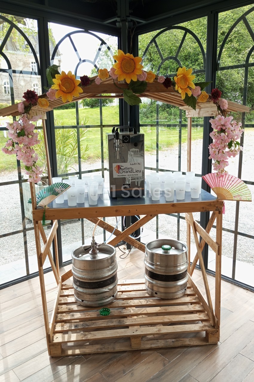 Stand en bois décoré de fleurs avec une tireuse à bière, des gobelets en plastique et des fûts dans un espace vitré.