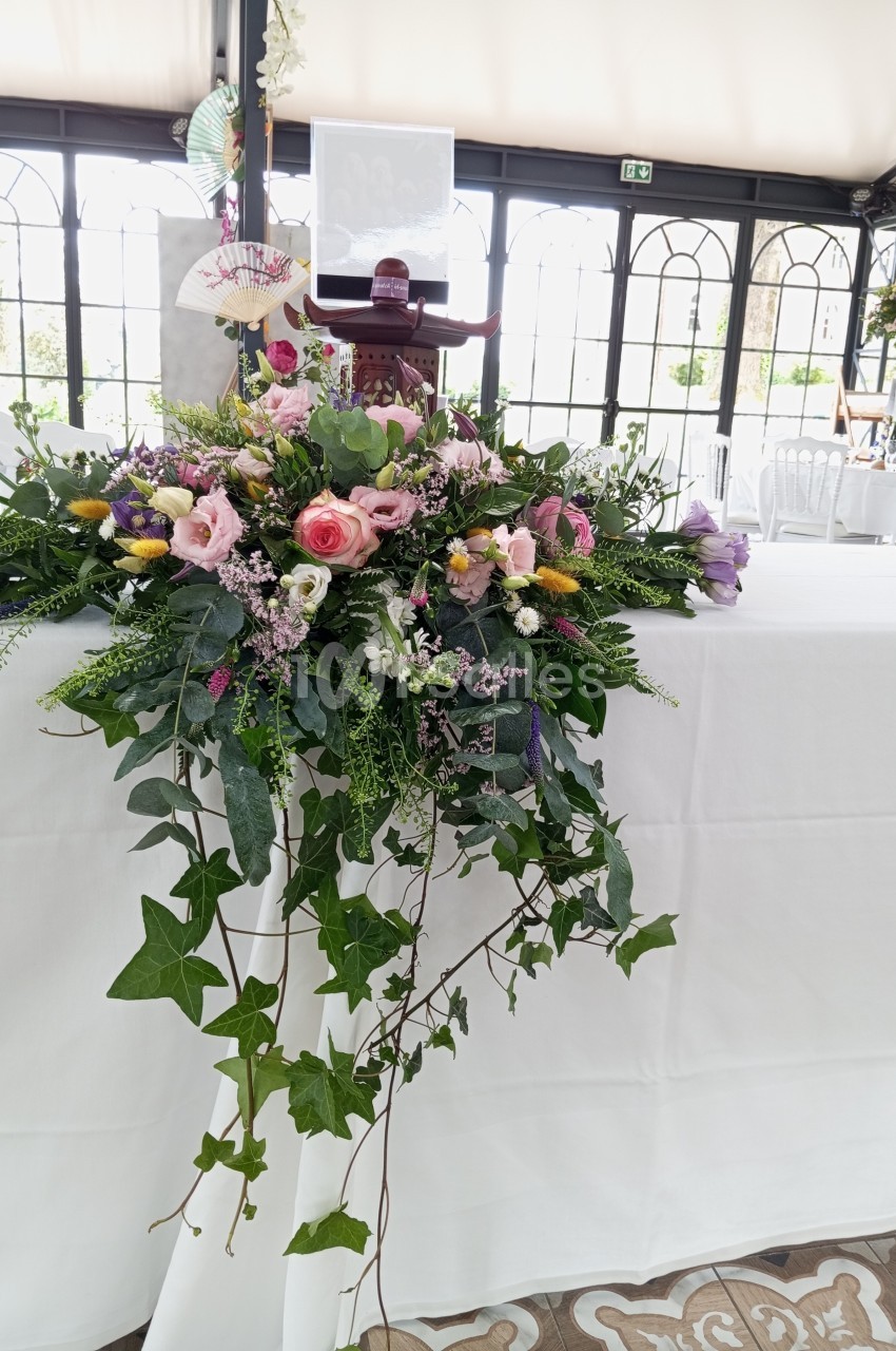 Arrangement floral coloré avec roses, feuillage et lierre, disposé sur une table blanche dans une salle lumineuse.
