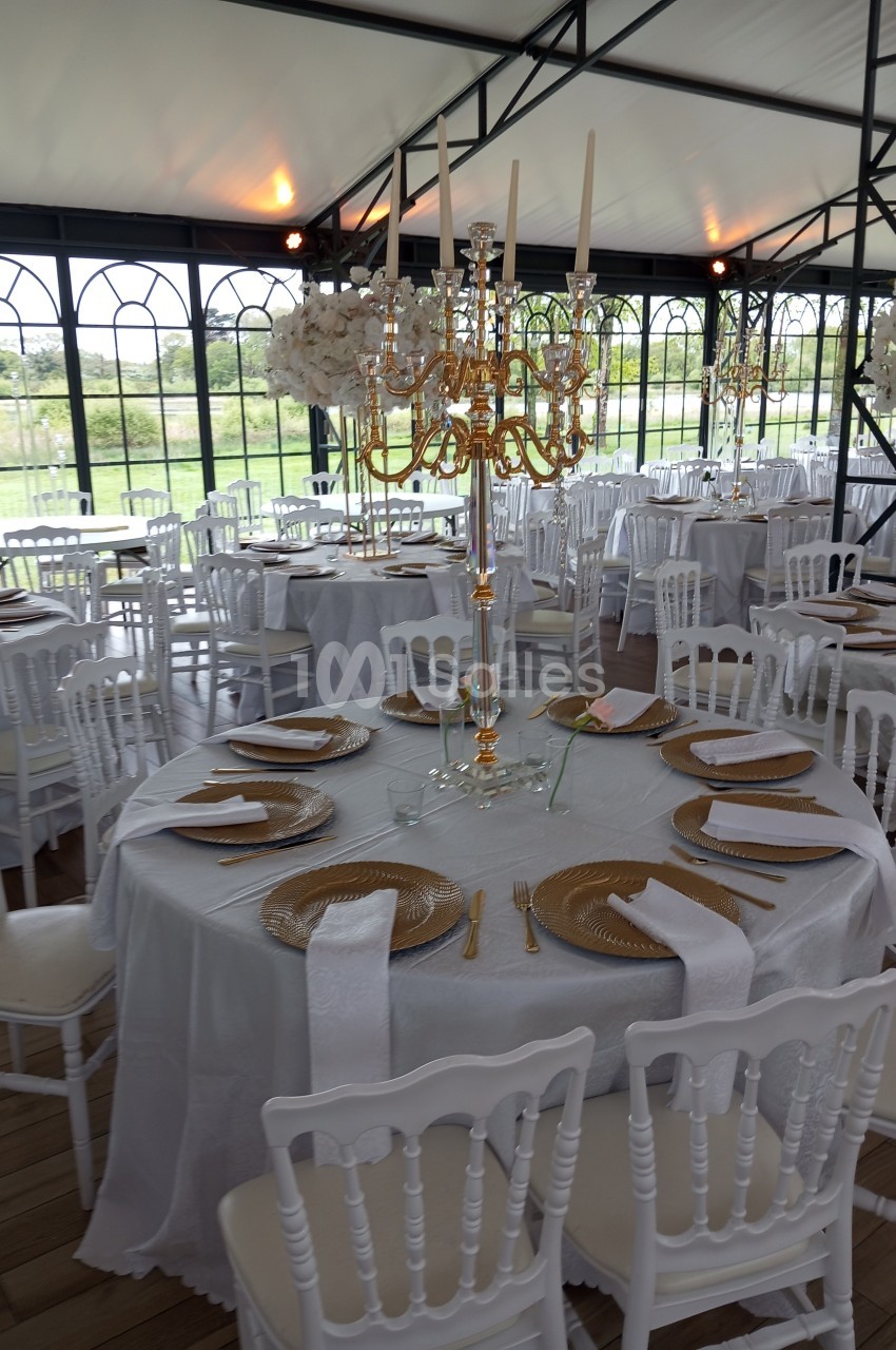 Salle de réception décorée avec des tables rondes dressées, nappes blanches, chandeliers dorés et chaises blanches.