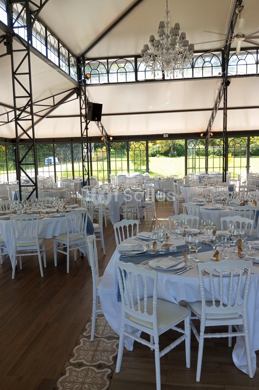 Salle de réception lumineuse avec tables rondes dressées, chaises blanches et vue sur un jardin à travers des baies vitrées.